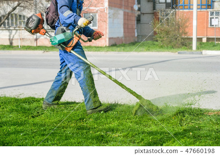 Man wearing overalls and rubber boots mowing green grass with gasoline lawn trimmer. Man wearing overalls and rubber boots mowing green grass with gasoline lawn trimmer. 47660198