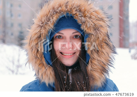 Young woman brunette looking at camera and smiling. Wearing blue down jacket with fur hood, face 47673619
