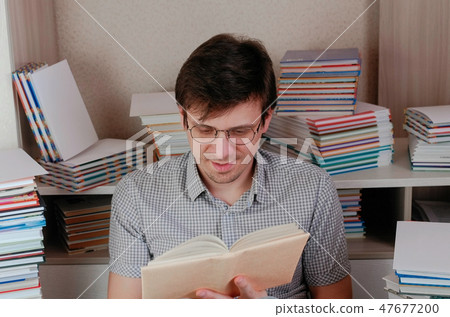 Young brunet man in glasses reads a book sitting among books. Young brunet man in glasses reads a book sitting among books. 47677200