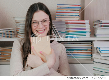 Young brunette woman in glasses laughs hugging a book sitting among books. Young brunette woman in glasses laughs hugging a book sitting among books. 47677295