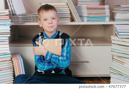 Seven-year-old boy hugging the book and smiling at the camera sitting among books 47677377