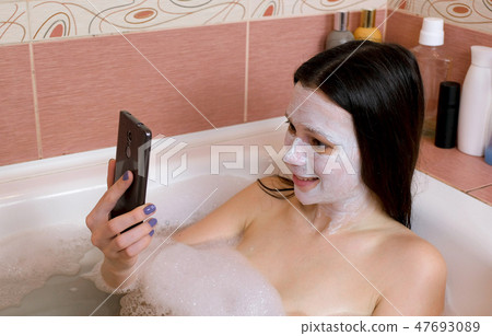 Brunette woman with a mask on her face lies in bathroom and chattin by mobile phone and laughing. Brunette woman with a mask on her face lies in bathroom and chattin by mobile phone and laughing. 47693089