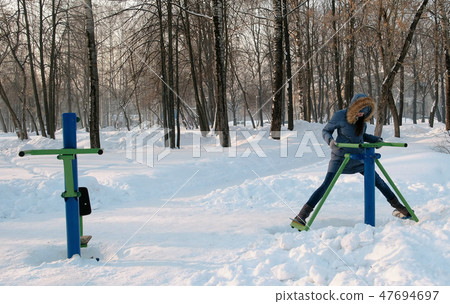 Woman in blue down jacket with hood is doing legs exercises on simulator in a winter Park Front view Woman in blue down jacket with hood is doing legs exercises on simulator in a winter Park Front view 47694697