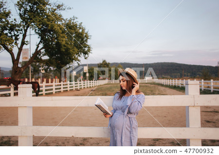Beautiful pregnant woman read the book on the farm. She stand by the fance and look in to the book 47700932