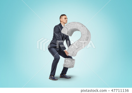 Businessman lifting up big stone question mark on blue background Businessman lifting up big stone question mark on blue background 47711136
