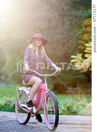 Blond fashionable long-haired attractive girl in short dress with lady bicycle in sunny park. 47713177
