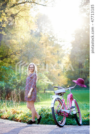 Blond fashionable long-haired attractive girl in short dress with lady bicycle in sunny park. 47713185