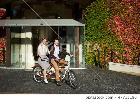 Young couple, handsome man and blond woman cycling tandem bike by building overgrown with red ivy. 47713186