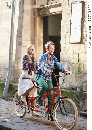 Young tourist couple, handsome man and pretty blond woman riding tandem bicycle along city street. 47713203