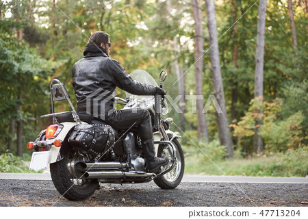 Bearded biker in black leather jacket on modern motorcycle on country roadside. 47713204