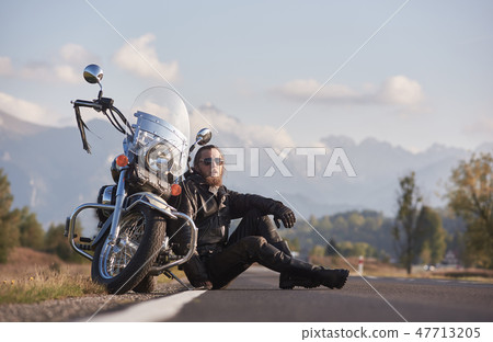 Athletic motorcyclist in black leather clothing sitting at modern powerful shiny motorbike. 47713205