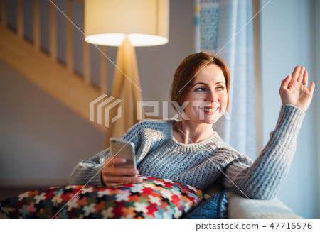 A young woman with smartphone sitting indoors on a sofa at home, waving. A young woman with smartphone sitting indoors on a sofa at home, waving. 47716576