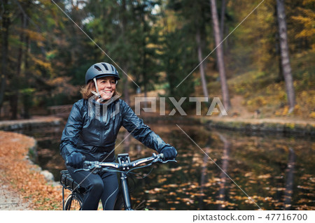 A senior woman with electrobike cycling outdoors on a road in park in autumn. A senior woman with electrobike cycling outdoors on a road in park in autumn. 47716700