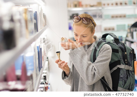 Blond young female traveler wearing travel backpack choosing perfume in airport duty free store. Blond young female traveler wearing travel backpack choosing perfume in airport duty free store. 47732740