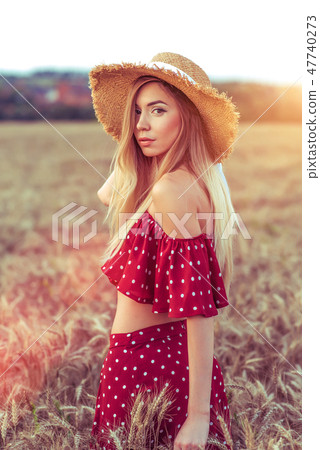 Girl in a wheat field in summer in red dress and straw hat. A woman walks across the field, long 47740273