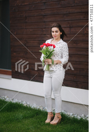 Beautiful girl with bouquet of red flowers stand by the house. Wooden background 47758811