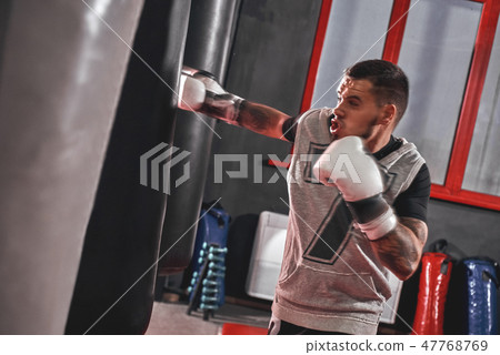 Pushing to the limit. Tattooed muscular athlete in sports clothing training hard on heavy punch bag 47768769