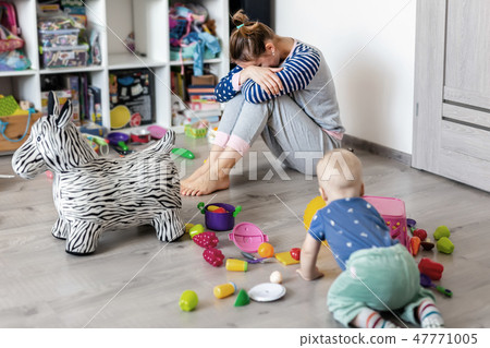 Tired of everyday household mother sitting on floor with hands on face. Kid playing in messy room 47771005