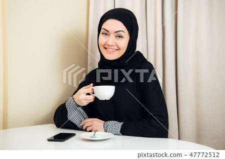 Arab women in hijab holding and drinking coffee cup sitting at home. 47772512