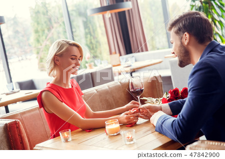 Young couple on date in restaurant sitting man giving small present box to woman happy Young couple on date in restaurant sitting man giving small present box to woman happy 47842900