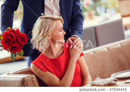 Young couple on date in restaurant man standing with bouquet behind sitting woman holding his hand 47844445