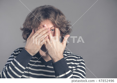 Handsome man with dark curly hair, bristle, hides face with hands, peeks through fingers, wears Handsome man with dark curly hair, bristle, hides face with hands, peeks through fingers, wears 47852496