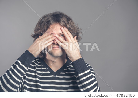 Portrait of young hipster man in sweater peeping at the camera through his fingers. Picture of Portrait of young hipster man in sweater peeping at the camera through his fingers. Picture of 47852499