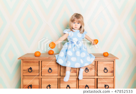 Toddler child baby girl in light blue dress seriously sitting on the wooden chest of drawers and Toddler child baby girl in light blue dress seriously sitting on the wooden chest of drawers and 47852533
