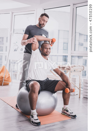Concentrated dark-skinned man sitting on the fitness ball 47858199