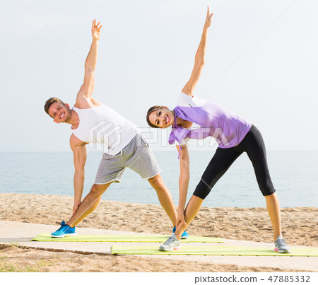 Smiling couple exercising yoga poses standing on beach Smiling couple exercising yoga poses standing on beach 47885332