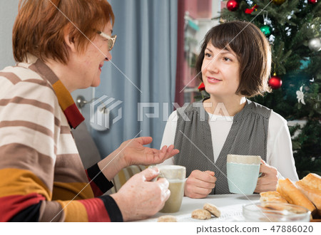 Two women drink tea and eat cakes near the New Year tree. Mother with daughter Two women drink tea and eat cakes near the New Year tree. Mother with daughter 47886020