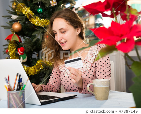 Happy young woman with credit card and laptop sitting at home against background of Christmas tree Happy young woman with credit card and laptop sitting at home against background of Christmas tree 47887149