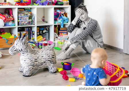 Tired of everyday household mother sitting on floor with hands on face. Kid playing in messy room Tired of everyday household mother sitting on floor with hands on face. Kid playing in messy room 47917719