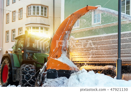 Big tractor with chains on wheel blowing snow from city street into dump truck body. Cleaning Big tractor with chains on wheel blowing snow from city street into dump truck body. Cleaning 47921130