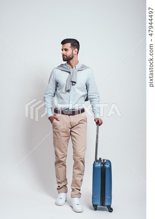 Ready to travel. Full length of handsome bearded young man with a suitcase keeping one hand in his Ready to travel. Full length of handsome bearded young man with a suitcase keeping one hand in his 47944497