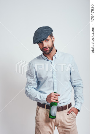 Bad boy. Bearded brutal man in hat holding a beer and looking ar camera while standing against grey Bad boy. Bearded brutal man in hat holding a beer and looking ar camera while standing against grey 47947980