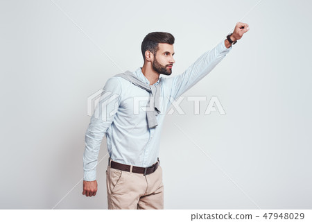 Superhero. Young bearded man is raising his hand up while standing against grey background Superhero. Young bearded man is raising his hand up while standing against grey background 47948029
