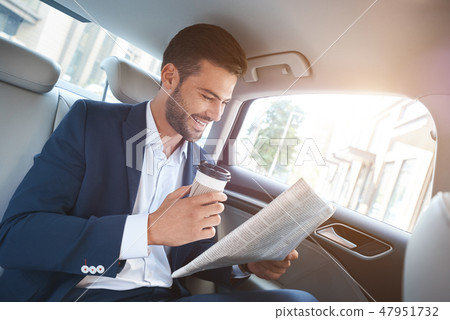 Young stylish businessman with newspaper drinking coffee on backseat of car and laughs Young stylish businessman with newspaper drinking coffee on backseat of car and laughs 47951732