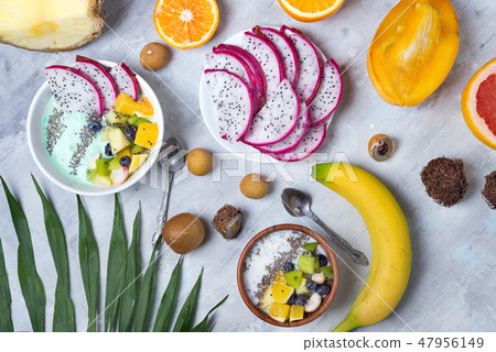 Breakfast table with yogurt acai bowls and fresh tropic fruits on a gray stone background with palm 47956149