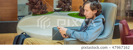 A man in the lounge area at the airport is waiting for his plane, uses a smartphone and headphones 47967162