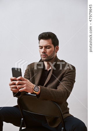 Social world. Stylish dark-haired man is using his phone while sitting on a chair isolated over 47979645