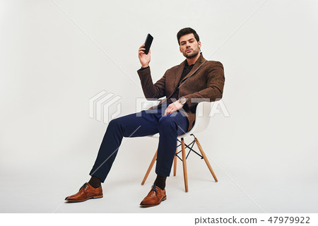 I have an idea! Stylish dark-haired man sitting on a chair, holding a phone in his hand and looking I have an idea! Stylish dark-haired man sitting on a chair, holding a phone in his hand and looking 47979922