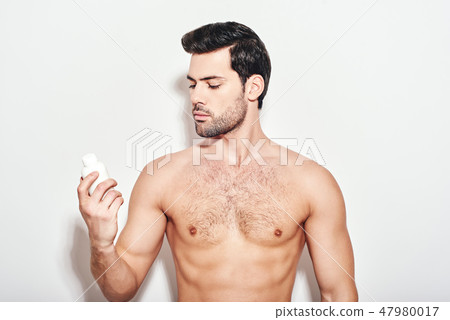 Confident in his skin. Handsome dark-haired man looks at the moisturizer that he uses standing 47980017