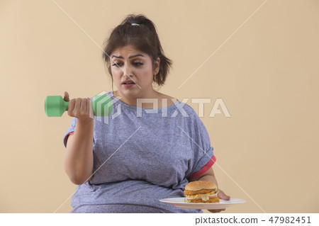 Sad Overweight Woman holding a burger and dumbbell Sad Overweight Woman holding a burger and dumbbell 47982451
