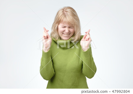Blonde woman wears green sweater, smiles broadly, keeps fingers crossed, hopes for good luck. Blonde woman wears green sweater, smiles broadly, keeps fingers crossed, hopes for good luck. 47992804