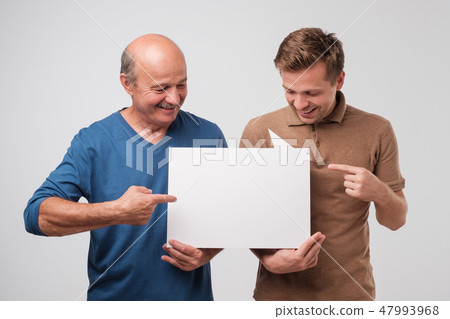 Two mature men father and son are holding a white empty billboard together Two mature men father and son are holding a white empty billboard together 47993968