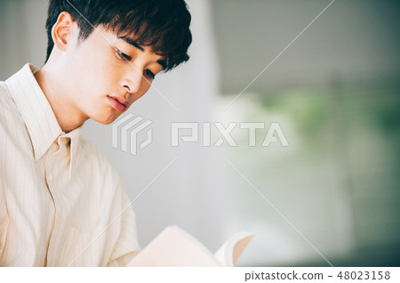 A man with a white shirt reading a book by the window 48023158