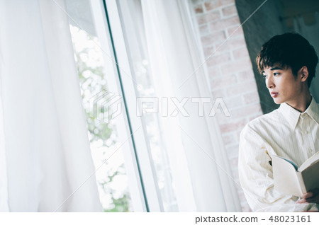 A man with a white shirt reading a book by the window 48023161
