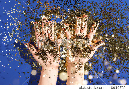 Hands of a child in gold glittering confetti on a blue background, top view. Birthday and holiday 48030731