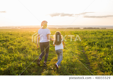 Rear view of mother and daughter running in green field with sunset on background Rear view of mother and daughter running in green field with sunset on background 48033329